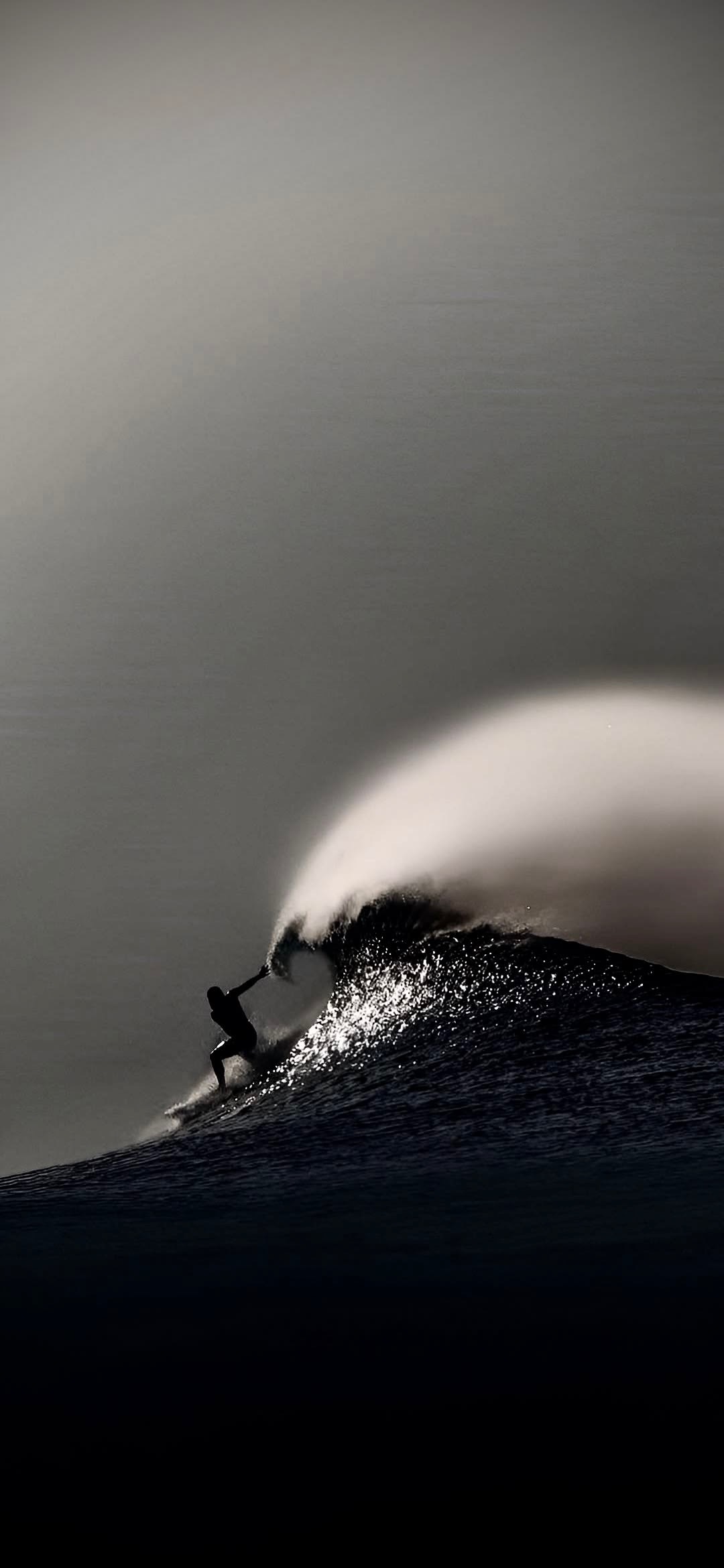 A lone surfer riding high on the face of a large wave photographed in black and white, spray lit from behind along the crest and the water falling away into darkness below.